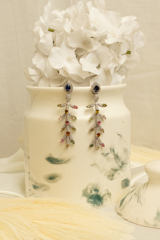 A pair of dangling statement earrings with silver-toned leaf-shaped charms set with clear stones and small multicolored gem accents, hanging from matching silver-toned hooks. The earrings are displayed on a white ceramic jar filled with white flowers, against a soft beige background.