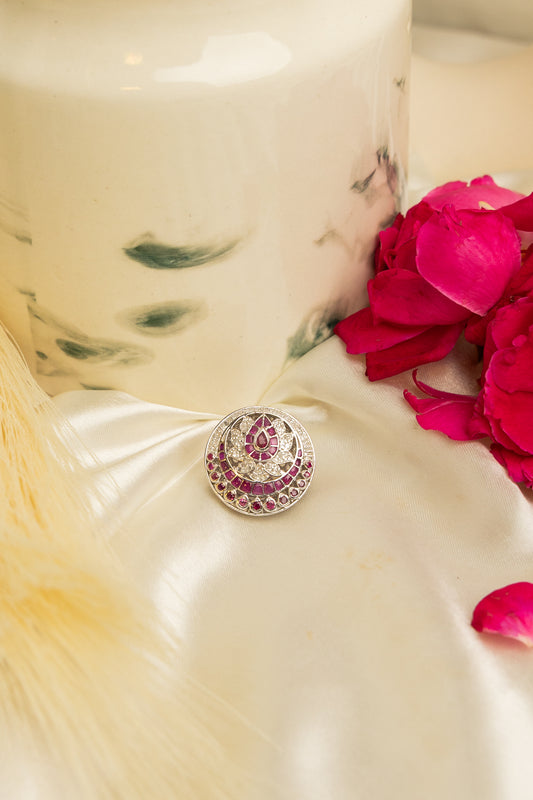 A close-up of a round silver-toned Rakhdi with a central pink gemstone and surrounding intricate pink and gold detailing, resting on draped cream satin fabric next to bright pink rose petals.